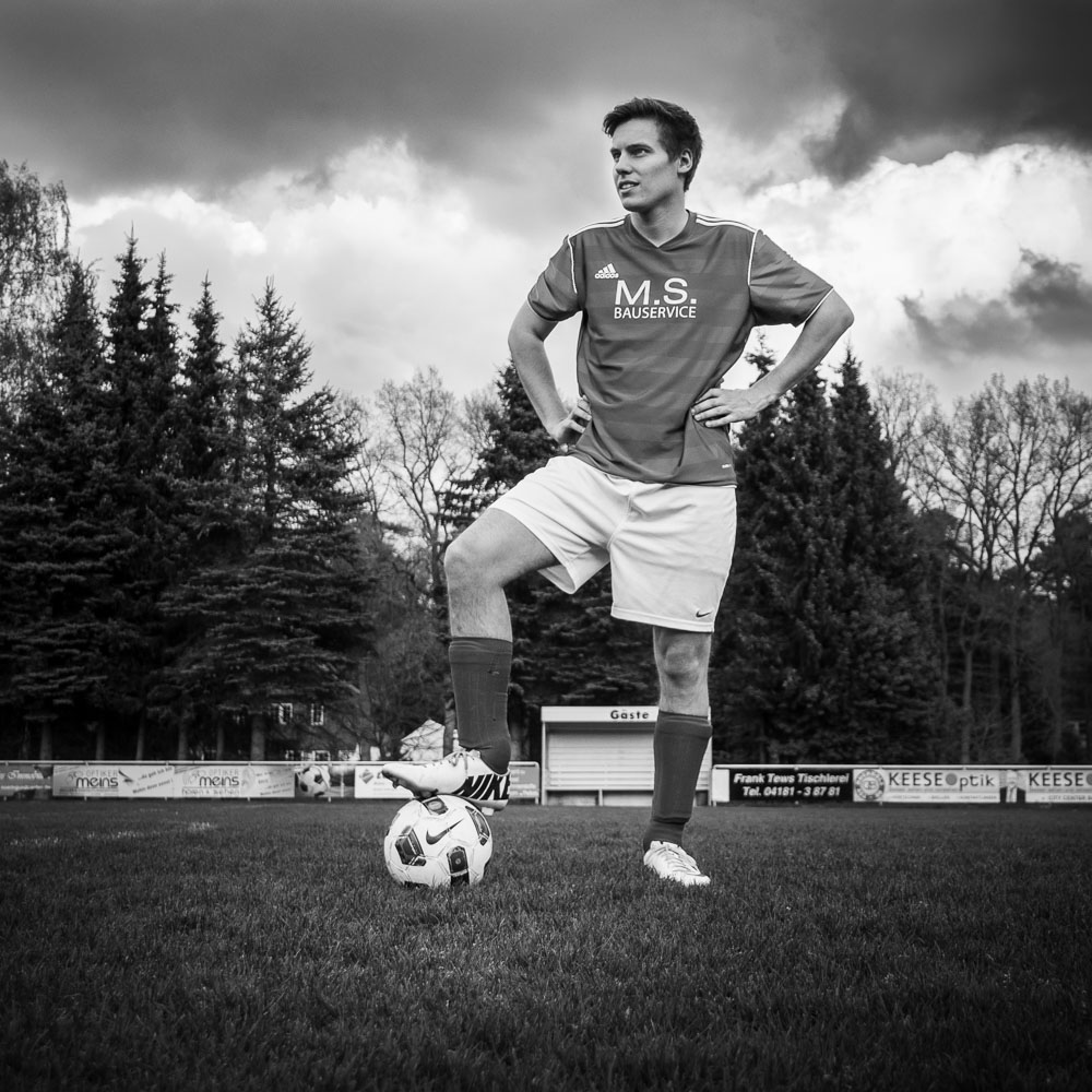 Schwarzweißes Sportlerportrait eines Fußballers, der mit einem Bein auf dem Ball steht und die Arme in die Hüfen stützt; Aufgenommen auf einem leeren Fußballplatz vor dramatischem Wolkenhimmel aus einer tiefen Perspektive.